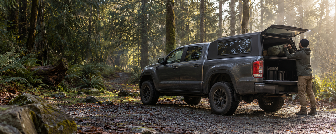 Camper loading gear into a pickup with a rigid shell at a forest campsite.